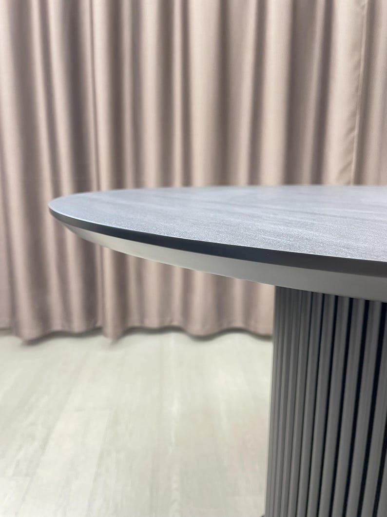 May include: A modern, round table with a gray, textured tabletop and a silver-colored edge. The table has a cylindrical base with vertical, gray, ridged detailing. The background features a neutral-colored curtain.