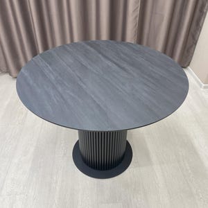 May include: A round, dark gray dining table with a textured surface. The table has a cylindrical base with vertical lines and a circular base. The table is approximately 48 inches in diameter.