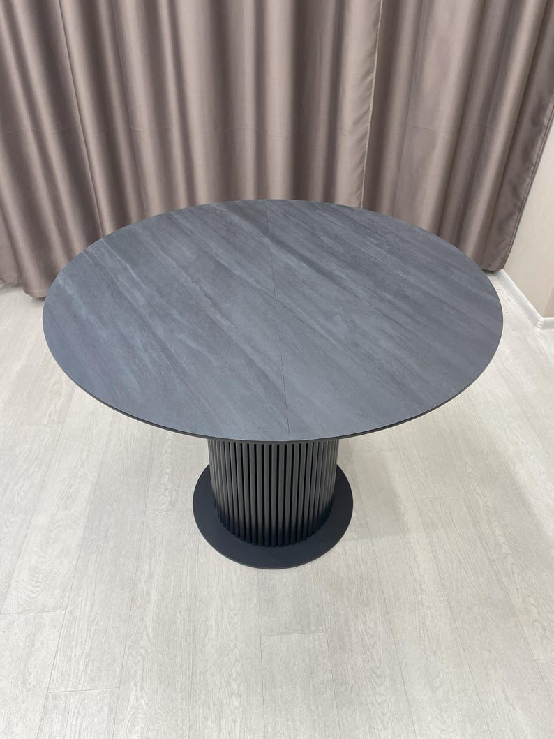 May include: A round, dark gray dining table with a textured surface. The table has a cylindrical base with vertical lines and a circular base. The table is approximately 48 inches in diameter.