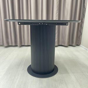 May include: A modern, dark gray round table with a cylindrical base. The base features vertical, ridged detailing. The tabletop is a flat, rectangular shape. The table is set against a neutral background.