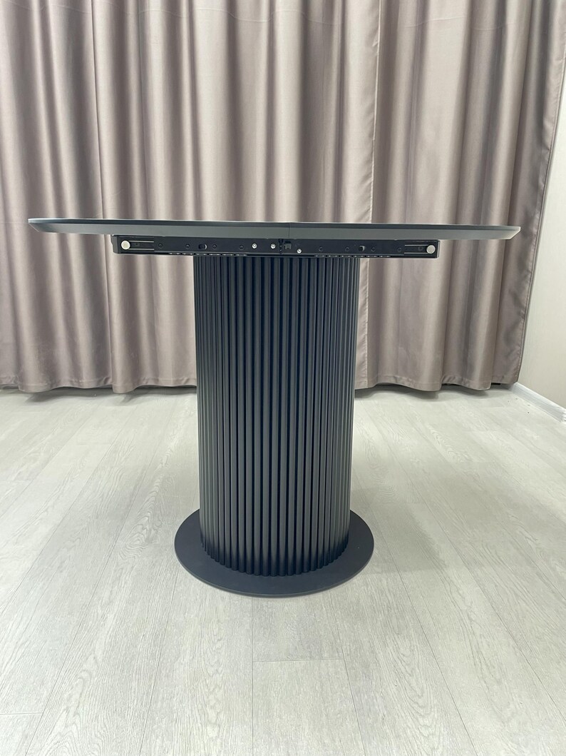 May include: A modern, dark gray round table with a cylindrical base. The base features vertical, ridged detailing. The tabletop is a flat, rectangular shape. The table is set against a neutral background.