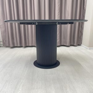 May include: A modern, round dining table with a dark gray tabletop and a cylindrical base. The base features vertical, grooved detailing and a circular foot. The table is set against a neutral-toned curtain and light-colored flooring.