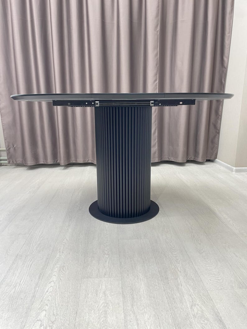 May include: A modern, round dining table with a dark gray tabletop and a cylindrical base. The base features vertical, grooved detailing and a circular foot. The table is set against a neutral-toned curtain and light-colored flooring.
