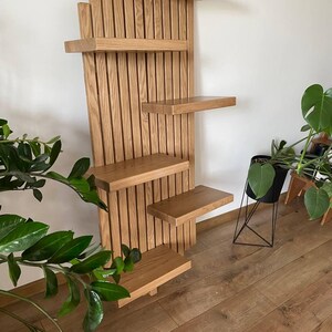May include: A wooden wall-mounted shelf unit with multiple shelves of varying sizes. The unit is made of light-colored wood and features a vertical slatted design. The shelves are arranged in an asymmetrical pattern, suitable for displaying plants or decor.