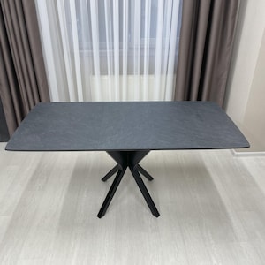 Op de afbeelding: Een zwarte rechthoekige eettafel met een leisteenachtig blad en een zwarte, stervormige basis. De tafel staat in een kamer met witte muren en een raam met witte gordijnen.