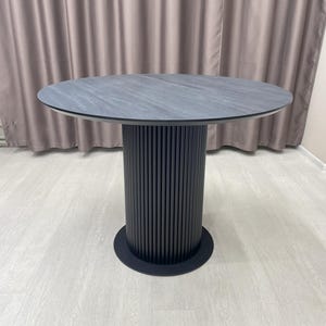 May include: A round dining table with a dark gray, stone-like top. The table has a black, cylindrical base with vertical grooves and a circular base. The table is set on a light-colored wood floor.