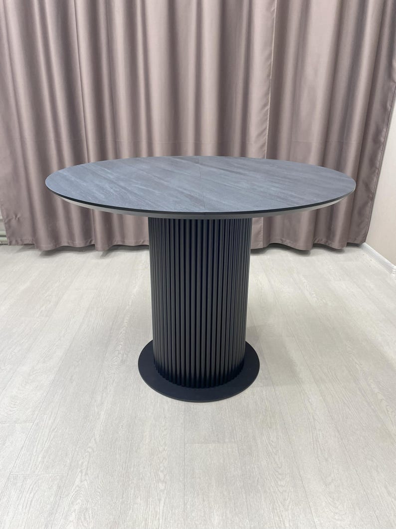 May include: A round dining table with a dark gray, stone-like top. The table has a black, cylindrical base with vertical grooves and a circular base. The table is set on a light-colored wood floor.