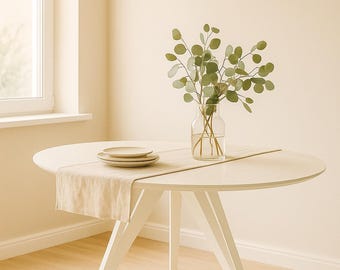 May include: A white round dining table with three legs, set with a beige table runner and two plates. A clear glass vase with green leaves sits on the table. A beige pendant light hangs above the table.