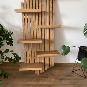 May include: A wooden wall shelf with a unique design. The shelf features multiple rectangular shelves of varying sizes, supported by vertical wooden slats. The wood has a natural, light brown color. The shelves are mounted on a white wall.