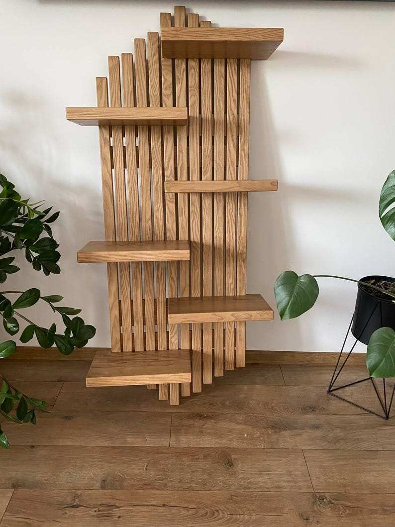 May include: A wooden wall shelf with a unique design. The shelf features multiple rectangular shelves of varying sizes, supported by vertical wooden slats. The wood has a natural, light brown color. The shelves are mounted on a white wall.