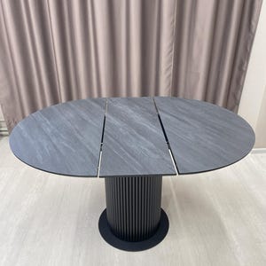 May include: A gray oval dining table with a textured surface. The table has a black cylindrical base with vertical lines and a round base. The table is approximately 60 inches in diameter.