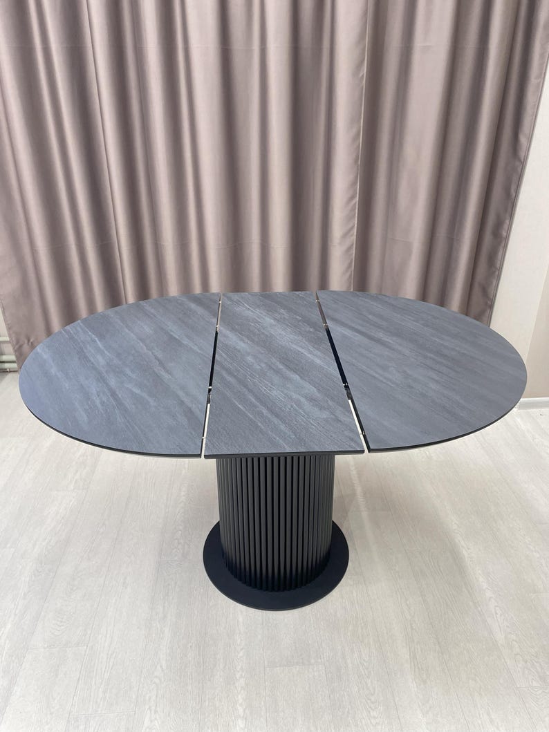 May include: A gray oval dining table with a textured surface. The table has a black cylindrical base with vertical lines and a round base. The table is approximately 60 inches in diameter.