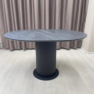 May include: An oval-shaped dining table with a dark gray stone-like top. The table has a cylindrical base with vertical grooves and a circular base. The table is a modern design.