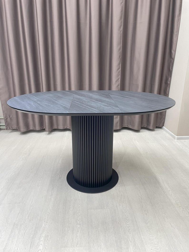 May include: An oval-shaped dining table with a dark gray stone-like top. The table has a cylindrical base with vertical grooves and a circular base. The table is a modern design.