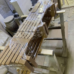 May include: A stack of wooden chairs, wrapped in clear plastic, ready for transport. The chairs are made of light-colored wood with a slatted back design. They are stacked on a metal stand in a workshop setting.