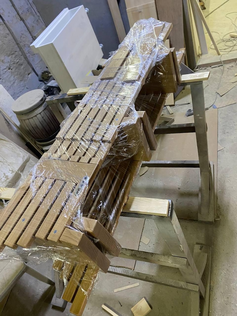 May include: A stack of wooden chairs, wrapped in clear plastic, ready for transport. The chairs are made of light-colored wood with a slatted back design. They are stacked on a metal stand in a workshop setting.