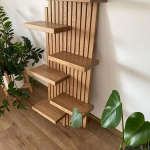 May include: A wooden wall shelf with four rectangular shelves. The shelf is made of light brown wood and has a vertical slatted back. The top of the shelf has a decorative, stepped design. The shelf is designed to hold plants or other decorative items.