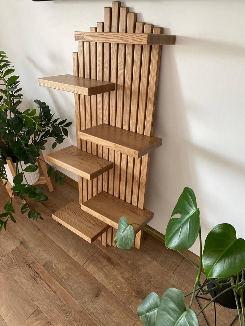 May include: A wooden wall shelf with four rectangular shelves. The shelf is made of light brown wood and has a vertical slatted back. The top of the shelf has a decorative, stepped design. The shelf is designed to hold plants or other decorative items.