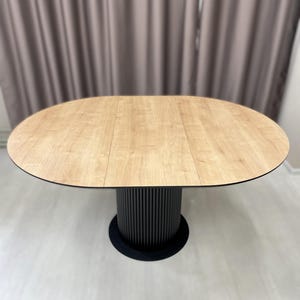 May include: Oval dining table with a light wood grain finish. The table has a black base with vertical ribbed detailing. The table is set against a neutral background.