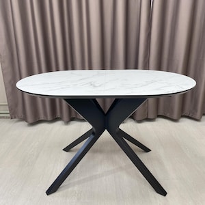 Peut inclure: Une table de salle à manger ovale avec un plateau blanc effet marbre et des pieds noirs inclinés. La surface de la table présente un motif veiné subtil. Les pieds sont d'une couleur sombre contrastante, créant une esthétique moderne.