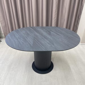 May include: Oval-shaped gray dining table with a textured surface. The table rests on a black, cylindrical base with vertical grooves. The base is supported by a circular black foot. The table is approximately 72 inches long.