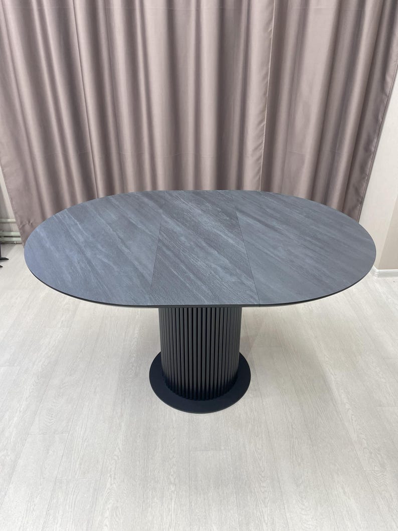 May include: Oval-shaped gray dining table with a textured surface. The table rests on a black, cylindrical base with vertical grooves. The base is supported by a circular black foot. The table is approximately 72 inches long.