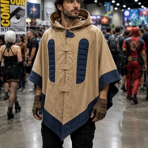 May include: A tan hooded poncho with navy blue trim and accents. The poncho has a button closure and is worn with brown gloves and black trousers. The background shows a convention hall with people and signage.