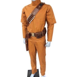 Może przedstawiać: Pomarańczowy kostium w całości, z koszulą z wysokim kołnierzem i spodniami, uzupełniony brązowym sk&oacute;rzanym paskiem i kieszeniami. Przez klatkę piersiową przebiega brązowy sk&oacute;rzany bandolier z białymi detalami w kształcie pocisk&oacute;w. W zestawie r&oacute;wnież kabura i cylindryczna sakwa.