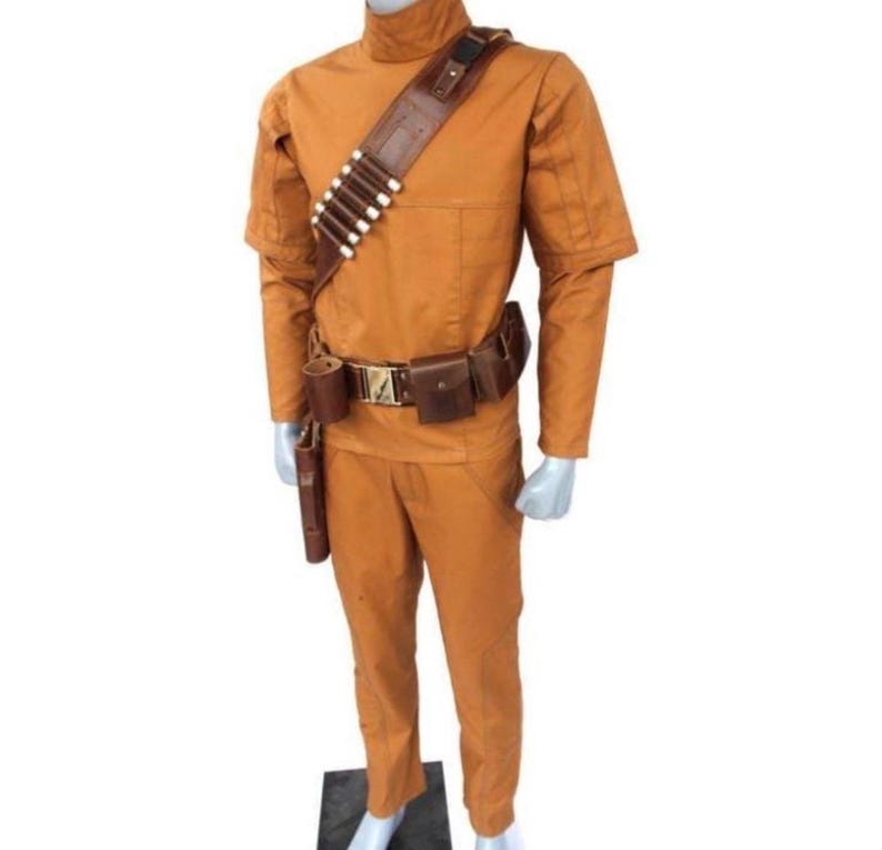 Może przedstawiać: Pomarańczowy kostium w całości, z koszulą z wysokim kołnierzem i spodniami, uzupełniony brązowym sk&oacute;rzanym paskiem i kieszeniami. Przez klatkę piersiową przebiega brązowy sk&oacute;rzany bandolier z białymi detalami w kształcie pocisk&oacute;w. W zestawie r&oacute;wnież kabura i cylindryczna sakwa.