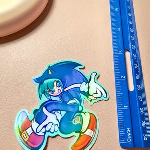 May include: A holographic sticker featuring an anime-style character wearing a blue Sonic the Hedgehog hoodie, positioned next to a blue ruler showing a height of approximately 3.5 inches. The character has teal hair, blue clothing, and red shoes.