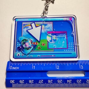 May include: A rectangular acrylic keyring featuring a digital interface graphic with a large white cursor, a music note icon on a disc, and a teal-haired character. A blue ruler placed underneath shows the keyring is approximately 7.6 cm wide.