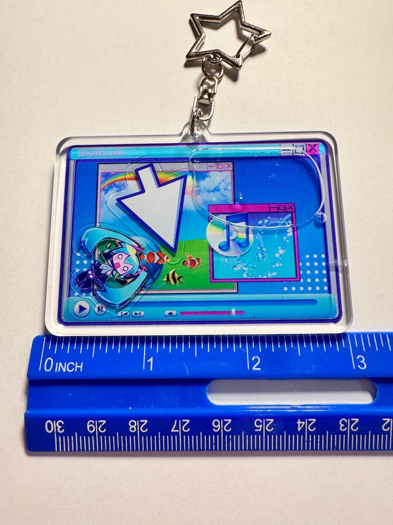 May include: A rectangular acrylic keyring featuring a digital interface graphic with a large white cursor, a music note icon on a disc, and a teal-haired character. A blue ruler placed underneath shows the keyring is approximately 7.6 cm wide.
