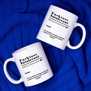 May include: Two white ceramic mugs with the text "Fucktose Intolerant" in black. The mugs have a definition that reads "The condition of being completely unable to tolerate other people's bullshit." The mugs are on a blue knitted surface.
