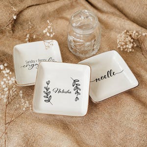 Op de afbeelding: Drie witte keramische ringenschalen met gouden rand. De schalen hebben verschillende ontwerpen en tekst. E&eacute;n schaal zegt "Sandra + Benny engaged", een andere zegt "Natasha" met een bloemmotief en de derde zegt "noelle" in cursief.