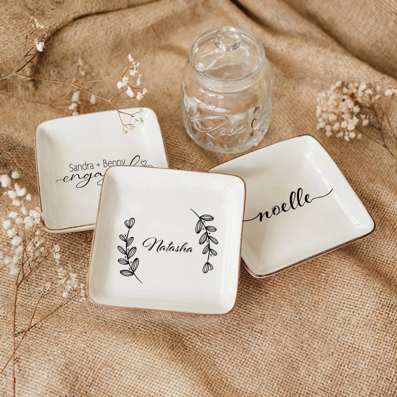 Op de afbeelding: Drie witte keramische ringenschalen met gouden rand. De schalen hebben verschillende ontwerpen en tekst. E&eacute;n schaal zegt "Sandra + Benny engaged", een andere zegt "Natasha" met een bloemmotief en de derde zegt "noelle" in cursief.