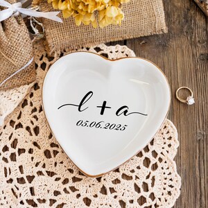 Op de afbeelding: Witte, hartvormige keramische ringenschotel met een gouden rand. De schotel heeft zwarte kalligrafie met de tekst "l + a 05.06.2025".