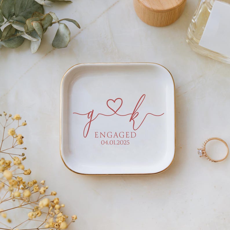 Op de afbeelding: Een witte keramische ringenschotel met een gouden rand. De schotel heeft een rood hart met de initialen "g" en "k" die met een lijn verbonden zijn. De tekst "ENGAGED 04.01.2025" is onder het hart gedrukt.