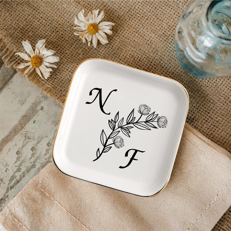 Op de afbeelding: Witte keramische sieradenbakje met een gouden rand. Het bakje heeft een zwart bloemenmotief met de letters "NF" in een handschrift.
