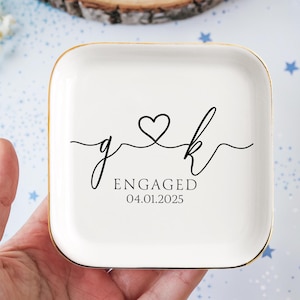 Op de afbeelding: Een witte keramische ringenschotel met een gouden rand. De schotel heeft een zwart ontwerp van twee initialen, "g" en "k", verbonden door een hart. De tekst "ENGAGED 04.01.2025" is onder de initialen gedrukt.