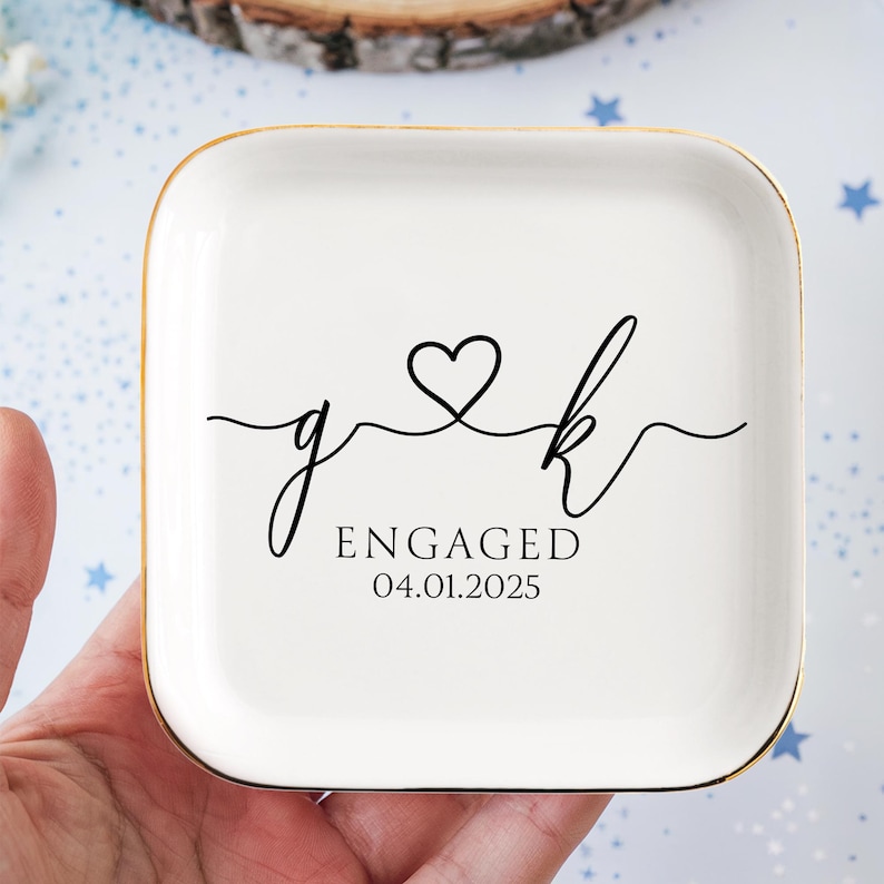 Op de afbeelding: Een witte keramische ringenschotel met een gouden rand. De schotel heeft een zwart ontwerp van twee initialen, "g" en "k", verbonden door een hart. De tekst "ENGAGED 04.01.2025" is onder de initialen gedrukt.