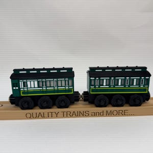 Könnte beinhalten: Zwei dunkelgrüne und schwarze Spielzeugeisenbahn-Personenwagen mit weißen Fensterdetails und gelben Zierlinien. Die Wagen sind miteinander verbunden und stehen auf einem Holzschienenstück, das den Schriftzug QUALITY TRAINS and MORE... eingraviert hat.