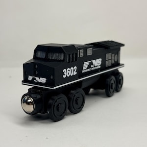 May include: A black toy train locomotive featuring the Norfolk Southern logo and the number 3602 printed in white on the side. The model includes black wheels with silver metallic centres and detailed cab windows, designed for use on wooden tracks.