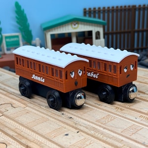 May include: Two wooden toy train cars, one labeled "Annie" and the other "Isabel". The cars are brown with white roofs and black wheels. Each car has a cartoon face. The cars are on a wooden track.