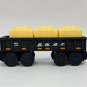 以下が含まれることがあります： 3つの黄色い貨物ブロックを積んだ黒いBNSF 3558のおもちゃの貨車。車両の両側には4つの黒い車輪があり、側面には白い文字でBNSFと記されています。デザインには小さなオレンジ色の長方形のアクセントが含まれています。