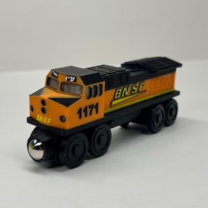 Könnte beinhalten: Eine Spielzeugeisenbahn-Lokomotive in Orange und Schwarz mit dem Text BNSF und der Nummer 1171 an der Seite. Die Lokomotive hat ein schwarzes Dach, schwarze Räder mit silbernen Zentren und ein Führerhaus mit Fenstern und runden Lichtdetails.