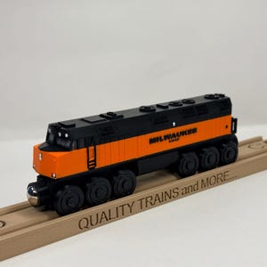 May include: A Milwaukee Road toy train engine featuring a black and orange colour scheme with black wheels. The train is positioned on a wooden track piece that has the text QUALITY TRAINS and MORE engraved on the side of the rail.
