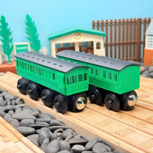 Può includere: Due vagoni di treno in legno verde con ruote nere e un paraurti anteriore argentato sono su binari di legno. I vagoni hanno un tetto grigio e fanno parte di una scena cittadina in miniatura.