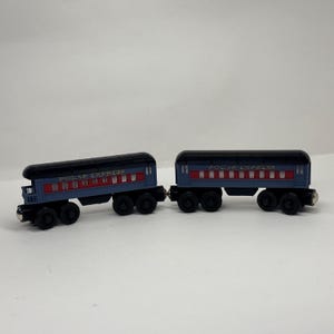 May include: Two blue and red toy train passenger cars with black roofs and wheels. Each car features the text POLAR EXPRESS printed in gold lettering along the side, with a series of small rectangular windows and four wheels on each side.
