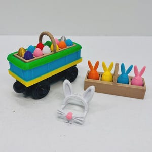 Puede incluir: Una composición temática de Pascua con un vagón azul y amarillo lleno de huevos pintados. Al lado, una caja de madera contiene cuatro figuras de conejitos en naranja, amarillo, azul y rosa. También hay una diadema de orejas de conejo blanca con una nariz rosa.