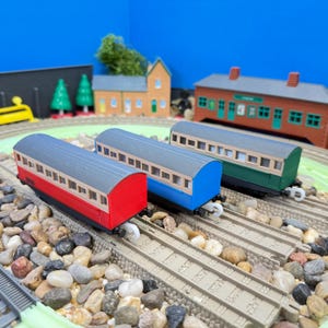 Può includere: Tre vagoni di treno giocattolo, rosso, blu e verde, sono su un binario modello. I vagoni hanno tetti grigi e telai di finestre beige. La scena include edifici e paesaggi in miniatura.